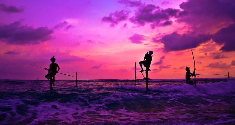 Silhouettierte Fischer auf Stelzen in der Abenddämmerung mit einem violetten Himmel.