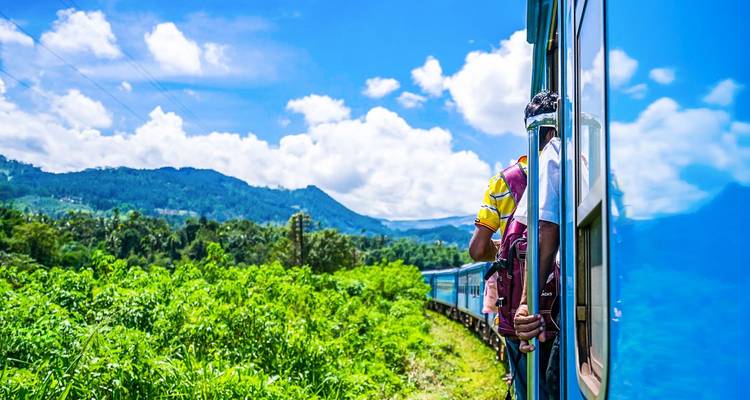 A traveler leans out of a bright blue train as it curves through lush green hills beneath a vivid blue sky.