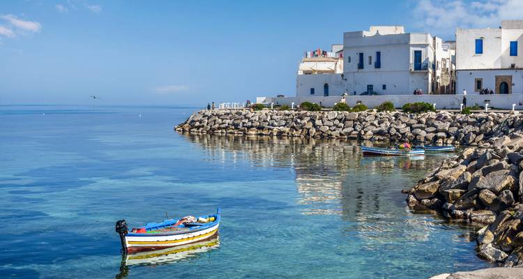 Buntes Fischerboot schwimmt auf kristallklarem türkisfarbenem Wasser neben weißgetünchten Häusern auf den Klippen eines mediterranen Dorfes.