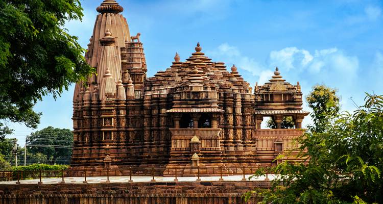 Ein kunstvoll geschnitzter Tempel von Khajuraho erhebt sich inmitten üppiger Vegetation unter blauem Himmel.