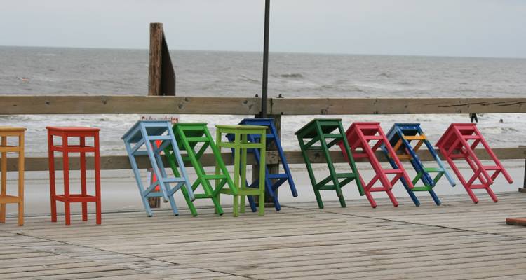Colorful wooden stools arranged on a boardwalk by a beach.