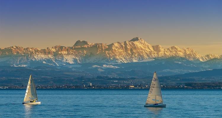 Two sailboats on a lake with snow-capped mountains in the background.