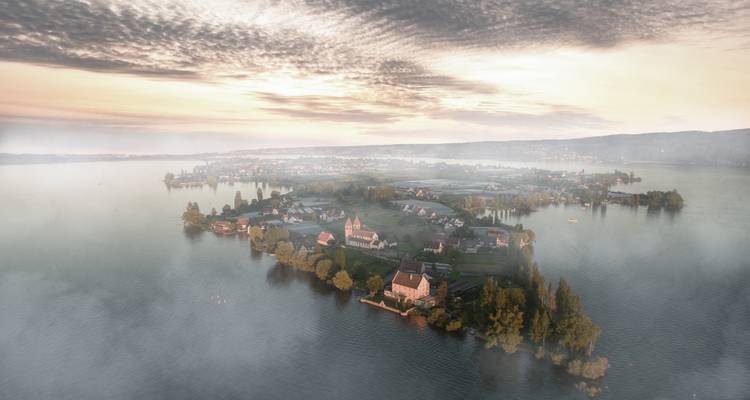 Island with structures surrounded by mist over a large expanse of water.