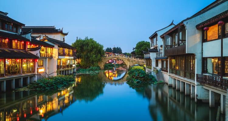 Peaceful canal town at twilight with arched stone bridge, lotus-filled water and traditional lit houses.