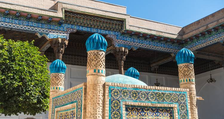 Verziertes gefliestes Mausoleum mit blauen Kuppeln und Backsteinpfeilern unter einem klaren blauen Himmel.