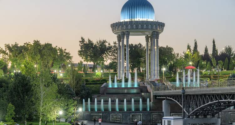 Modernes Denkmal mit blau gefliester Kuppel, Säulen und beleuchteten Springbrunnen in der Abenddämmerung in einem angelegten Park.