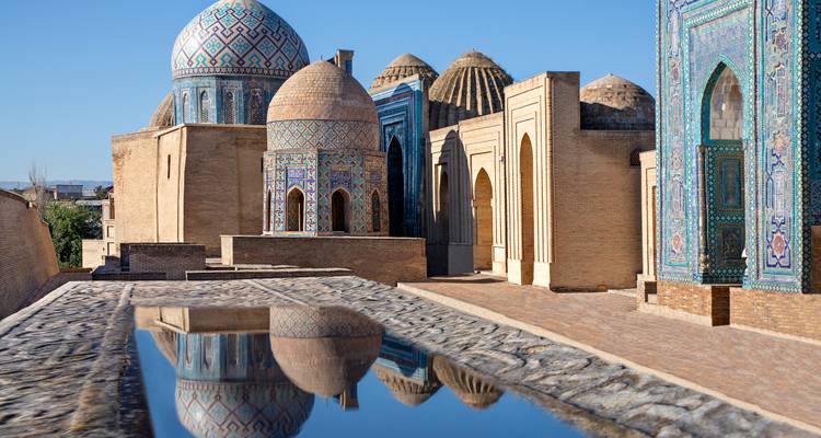 Reihe von blau gefliesten Kuppeln und Ziegelmausoleen, die sich in einem stillen Wasserkanal in Samarkand spiegeln.
