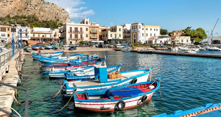 Rij van helderblauwe vissersboten afgemeerd in een pittoreske Siciliaanse haven met dorpsdecor op de achtergrond.