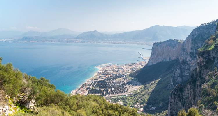 Breed panoramisch uitzicht over een turquoise baai en kuststad omlijst door dramatische kliffen en verre bergen.