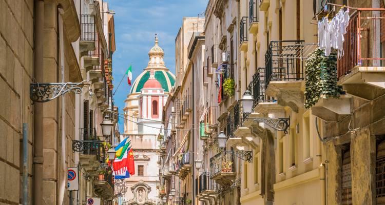 Schilderachtige Italiaanse steeg met balkons, vlaggen en een kleurrijke koepelkerk aan het einde van de straat onder blauwe luchten.