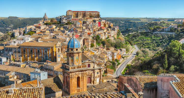 Verhoogd uitzicht over de honingkleurige barokstad geclusterd op een heuvel met een blauw betegelde kerkkoepel op de voorgrond.