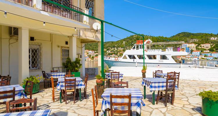 Taverne im Freien mit blau karierten Tischdecken mit Blick auf ein angedocktes Ausflugsboot an einem sonnigen Tag.