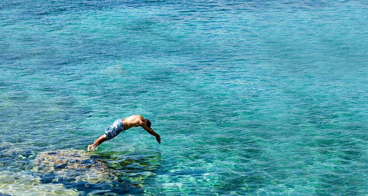Ein Mann springt von einem Küstenfelsen in funkelndes türkisfarbenes Meerwasser.