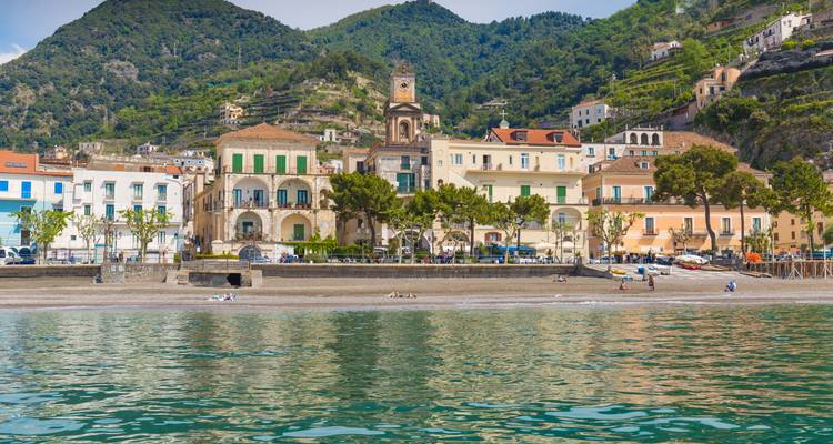 Pastellfarbene Gebäude an der Strandpromenade und ein Glockenturm säumen einen kleinen Strand an der Amalfiküste mit glitzerndem grünem Wasser im Vordergrund.