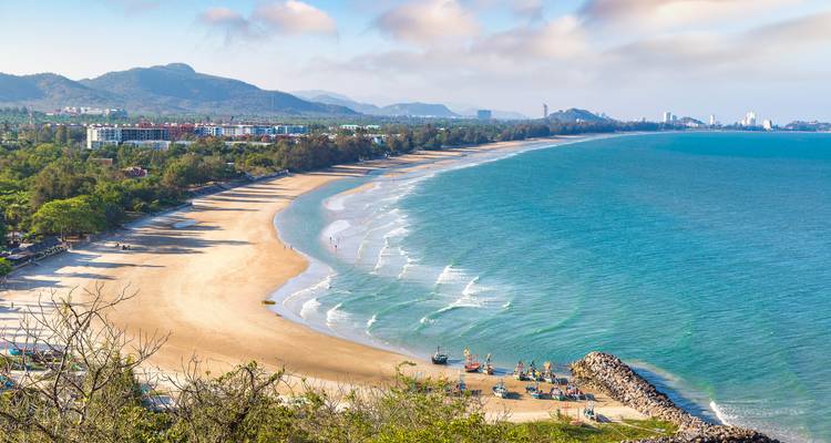 Lange geschwungene goldene Sandstrände und sanfte Brandung erstrecken sich entlang Hua Hins Küste mit bewaldeten Hügeln und fernen Resorts.