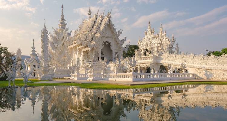 Die kunstvollen weißen Türme und Skulpturen von Wat Rong Khun spiegeln sich perfekt in einem ruhigen Teich unter sanftem Nachmittagslicht wider.