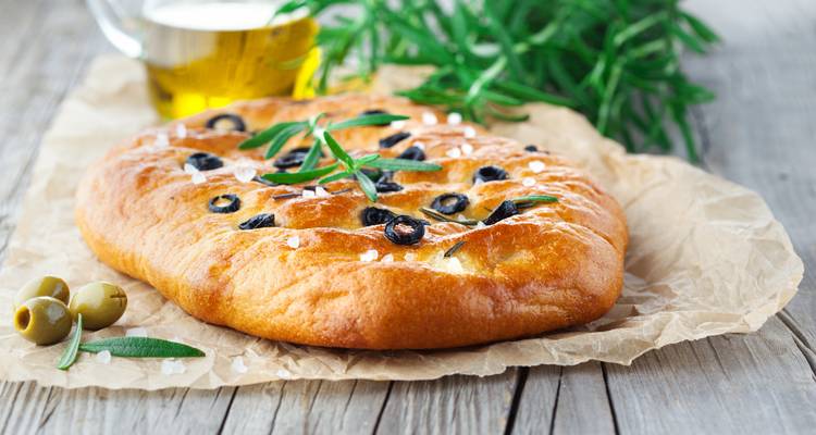 Frisch gebackene Focaccia belegt mit Oliven, Rosmarin und Meersalz, ruhend auf Pergamentpapier mit Olivenöl im Hintergrund.