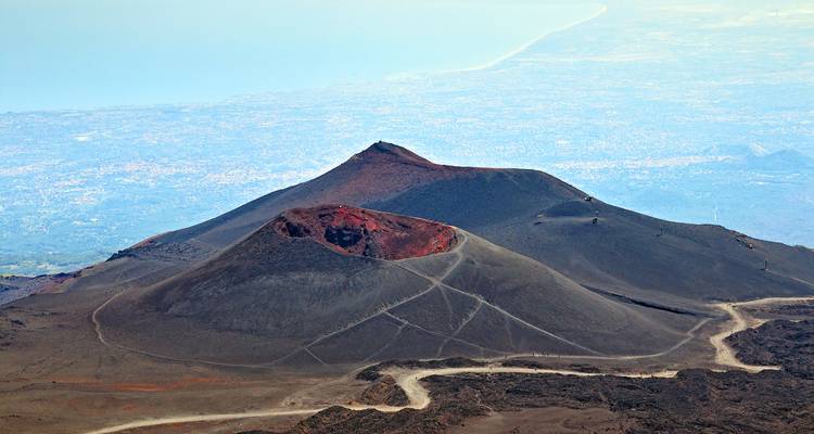 Eine Luftaufnahme der schwarzen Vulkankrater des Ätna mit sich schlängelnden Pfaden und der fernen sizilianischen Küste.