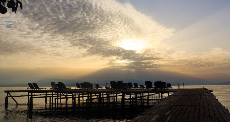 Muelle de madera bordeado de tumbonas vacías se extiende sobre aguas tranquilas mientras el sol sale detrás de nubes dramáticas.