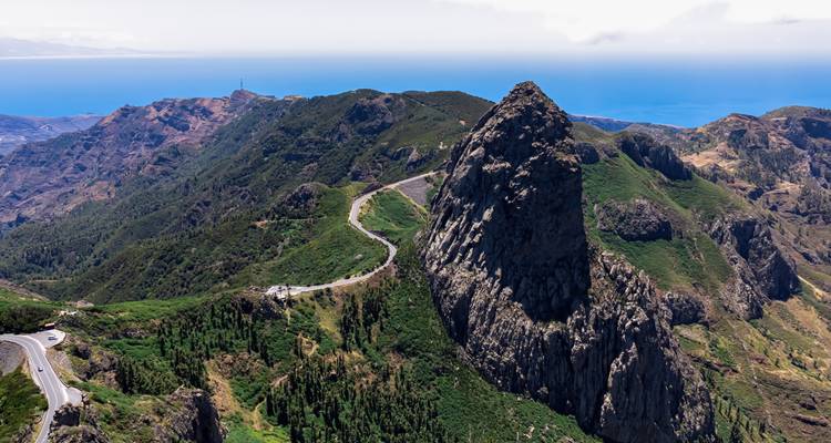 Majestueuze vulkanische monoliet rijst op naast een kronkelende bergweg met weids zeezicht in de verte.