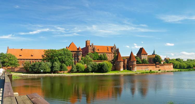 Weitläufige Ansicht der Marienburg, die sich an einem klaren Tag in einem ruhigen Fluss spiegelt.
