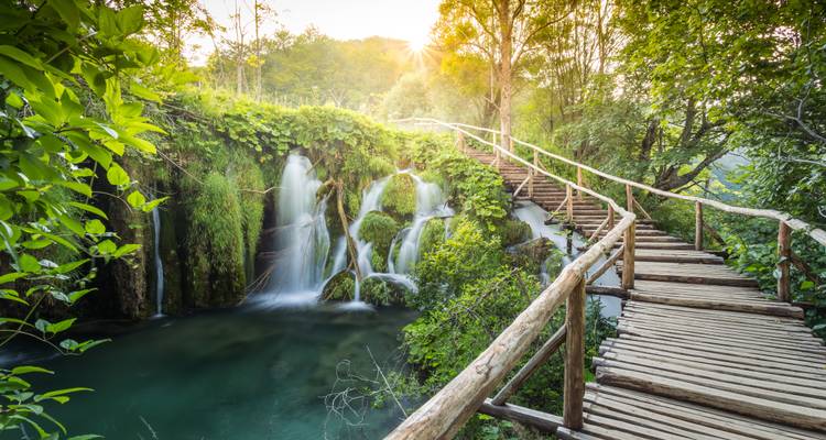 Boardwalk beside a tranquil waterfall and turquoise pool framed by lush vegetation in Plitvice Lakes National Park
