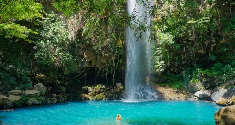 Persona nadando en una piscina turquesa al pie de una cascada.