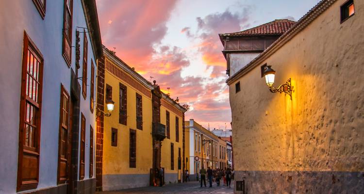 Narrow old town alley at dusk with warm street lights and people strolling beneath a vivid sunset sky