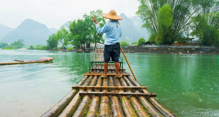 Floßfahrer mit Strohhut stakt ein Bambusfloß entlang eines jadegrünen Flusses mit nebligen Karstgipfeln im Hintergrund