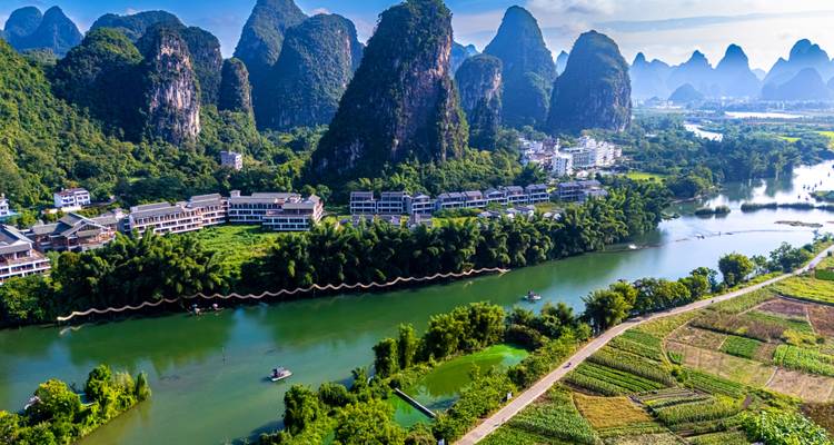 Luftpanorama einer Flussstadt, eingebettet zwischen hoch aufragenden Kalkstein-Karstgipfeln und üppigen Feldern