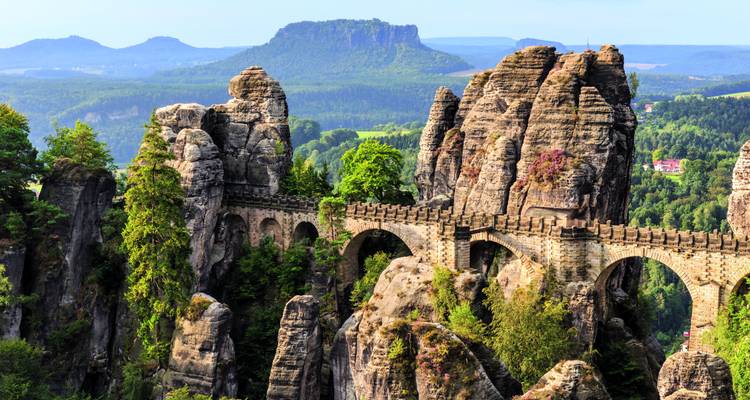 Le pont Bastei emblématique s'étend sur des piliers de grès spectaculaires avec des collines boisées et des mesas au loin.