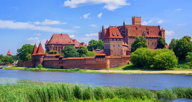 El majestuoso castillo de Malbork de ladrillo rojo con torres defensivas junto a un río tranquilo en un día soleado.