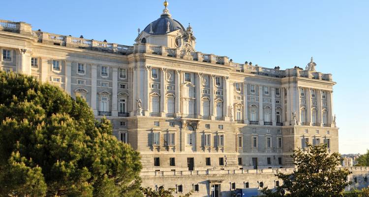 Großer Königspalast von Madrid im warmen Nachmittagslicht mit üppigen Bäumen im Vordergrund.