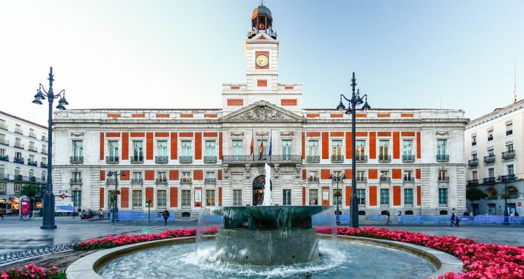 Puerta del Sol mit historischem Regierungsgebäude mit roten Verzierungen und zentralem Springbrunnen, umrahmt von rosa Blüten.
