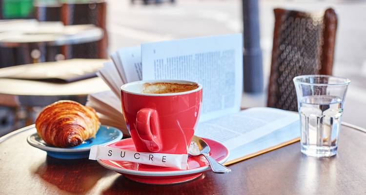 Pariser Café-Tisch mit einer roten Cappuccino-Tasse, Croissant, aufgeschlagenem Buch und Zuckerpackung mit der Aufschrift „SUCRE".