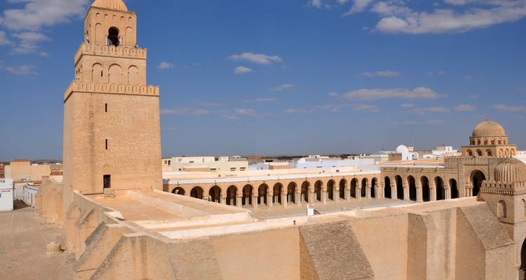 Wijde blik over de Grote Moskee van Kairouan met zijn vierkante minaret, arcadebinnenplaats en omringende lage witte gebouwen onder een blauwe hemel met verspreide wolken.