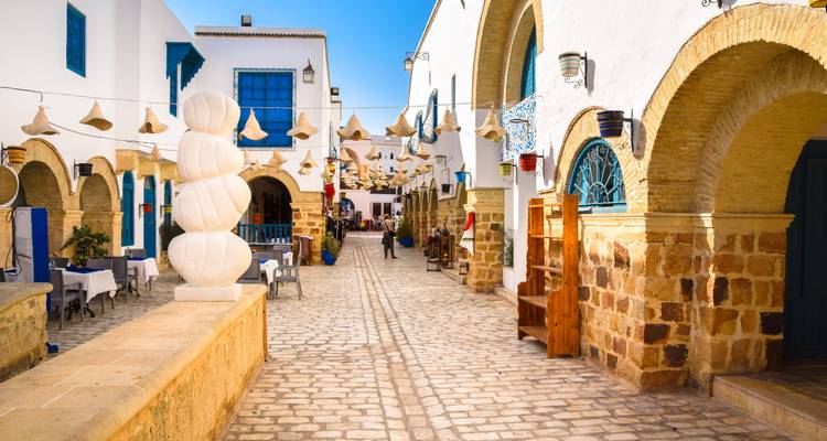 Charmant witgekalkt steegje met blauw omrande deuren, hangende stroolampen en een eenzame voetganger die over de geplaveide straat loopt.