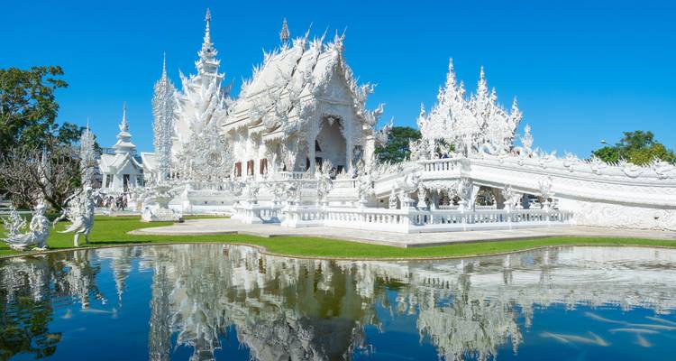 Strahlend weißer Wat Rong Khun Tempel, der sich in einem ruhigen Teich unter einem klaren blauen Himmel spiegelt.