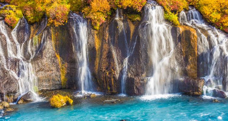Türkisfarbenes Wasser, das über mehrere enge Wasserfälle über dunkles Lavagestein fließt und von Herbstlaub umrahmt wird.