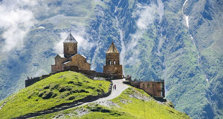 Besucher, die den Weg zur Gergeti-Dreifaltigkeitskirche auf einem üppigen Hügel mit nebligen Bergen hinaufgehen.