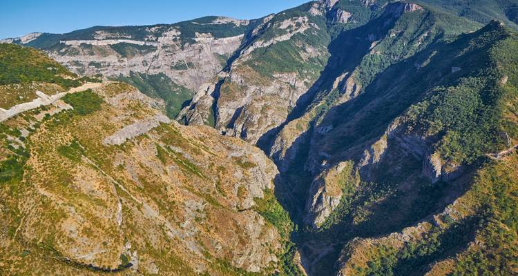 Steile zerklüftete Schlucht mit geschichteten Felswänden und spärlicher Vegetation unter klarem blauen Himmel.