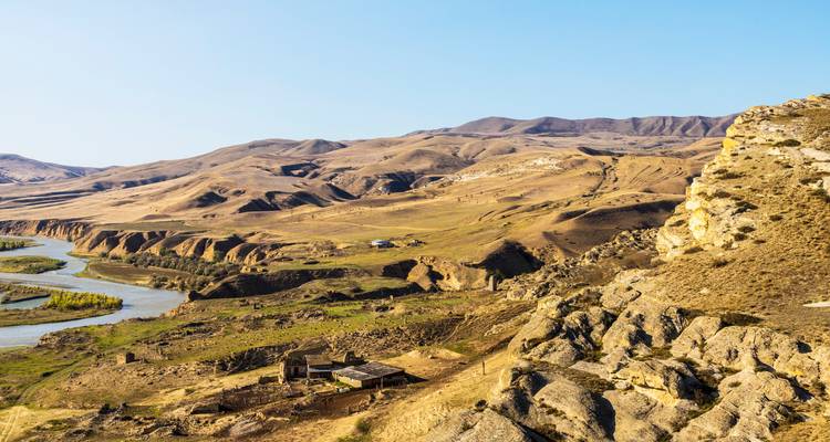Trockene wellige Hügel und Flusstal mit kleinen verlassenen Steinbauten in trockener Landschaft.