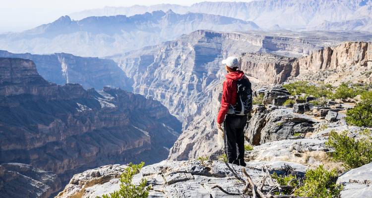Excursionista con chaqueta roja se encuentra en el borde rocoso contemplando el vasto cañón de Jebel Shams.