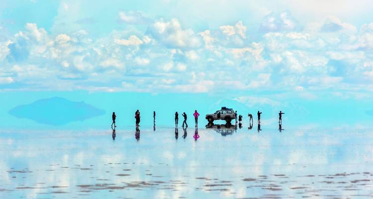 Menschen stehen auf einer spiegelnden Salzebene mit einem Fahrzeug in der Ferne.
