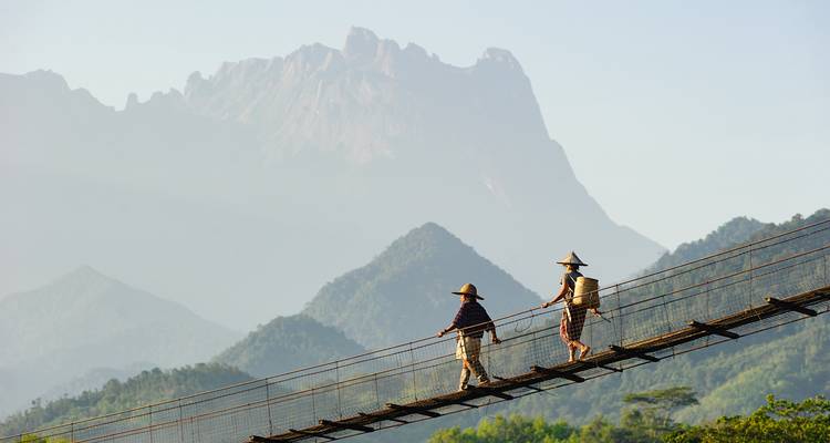 Zwei Wanderer mit kegelförmigen Hüten überqueren eine schmale Hängebrücke mit nebligen, zerklüfteten Bergen im Hintergrund