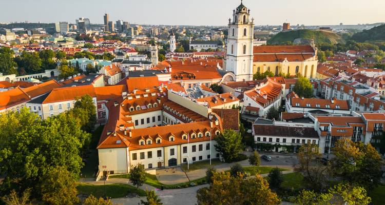 Luftaufnahme der Altstadt von Vilnius mit roten Ziegeldächern, Kirchtürmen und der umliegenden Stadtlandschaft zur goldenen Stunde.