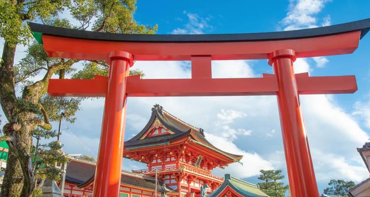 Vibrante puerta torii roja se alza ante un tejado de santuario sintoísta bajo un cielo azul brillante