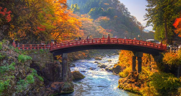 Un puente rojo histórico atraviesa un río rocoso rodeado de bosques otoñales bajo una cálida luz solar