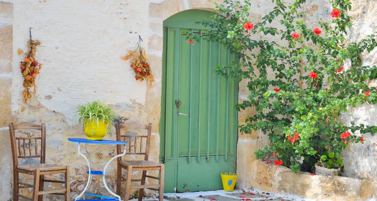 Charmante cour intérieure avec porte verte, table et chaises bleues, et hibiscus en fleurs contre un mur blanc.