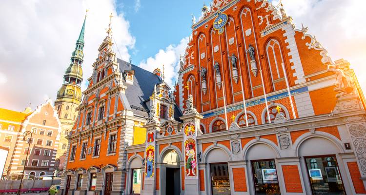 Verziertes rotes Backsteinhaus der Schwarzhäupter und Peterskirchturm in Riga, beleuchtet von warmer Sonne und blauem Himmel.